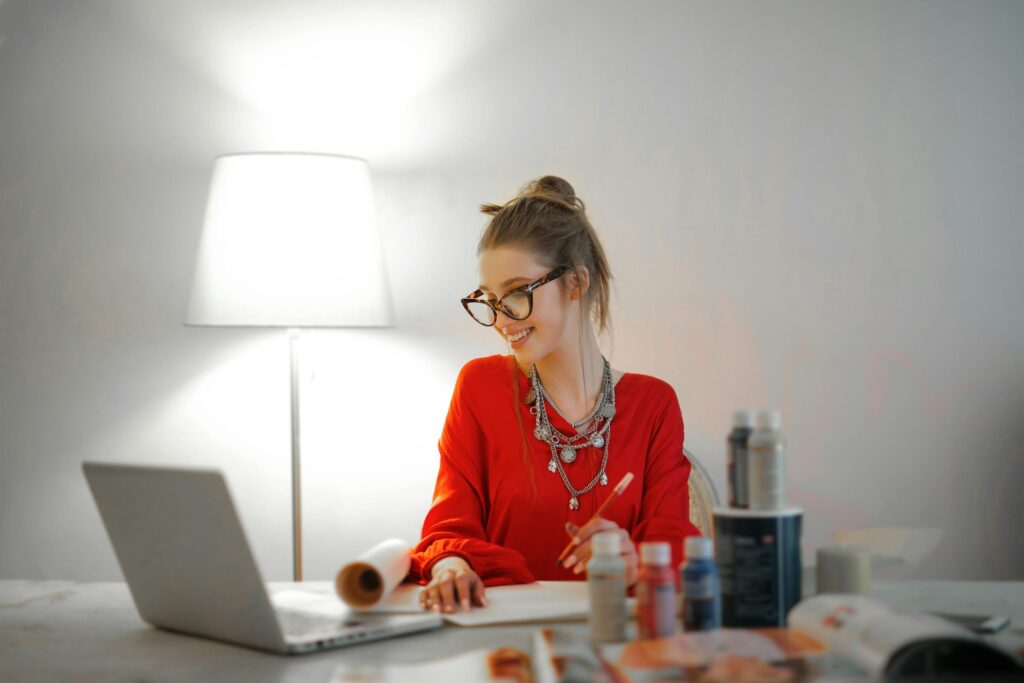 Smiling woman in red using laptop and art supplies at home office desk.